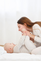 Happy cheerful young mom smiling playing with her little baby boy lying in bed at home. family portrait, white clothes, light house interior