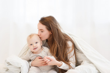Fototapeta na wymiar happy young mother kisses her little baby boy gently before bedtime. Mather's love and care. White clothes, light interior