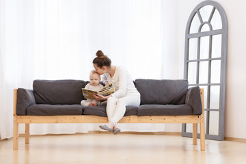 Pretty young mother reading a book to her little baby boy on a sofa in a light bedroom of house. Family holiday and togetherness.