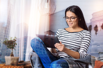 Fototapeta premium Convenient device. Curious lovely young woman sitting in a comfortable armchair and looking satisfied while trying to use her new modern tablet