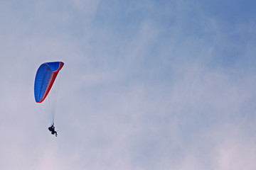 Pilote de parapente volant sur un aile bleue et rouge dans un ciel nuageux de Savoie en France.	