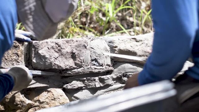 Two men using tuckpointing trowels to press mortar into the rocks in in a stone wall being built.