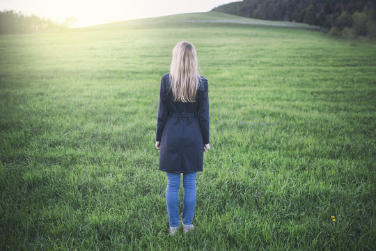 Back View Of Blonde Woman Standing On The Sunny Meadow.  