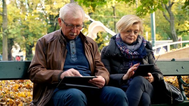 Mature couple using laptop and tablet computer sitting on a bench in the city park
