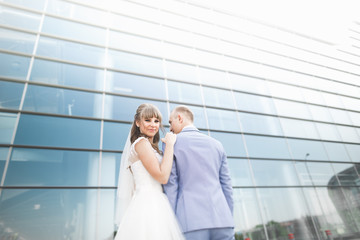 Lovely happy wedding couple, bride with long white dress posing near modern building