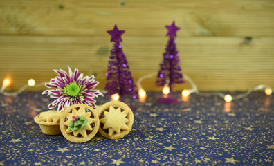 warm christmas food photography picture of traditional mince pies with fresh cut purple winter flower with tree decorations and fairy lights in background on star table cover