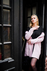 Blonde girl at glasses and pink coat, black tunic posed against wooden doors.