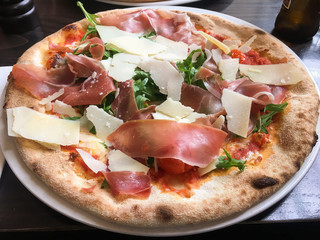 Fresh tasty pizza with a crust on it is on a white plate on a wooden table. Ingredients of pizza: green salad of wild rocket, Parmesan cheese and ham. Concept: favorite and popular food in the world.