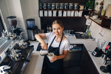 Portrait of smiling young Caucasian woman pouring hot milk in coffee. Waitress holding white mug cup in cafe. Person at work, small business concept. View from top above