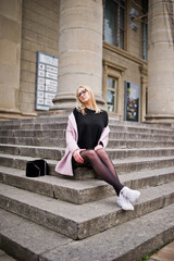 Fototapeta na wymiar Blonde girl at glasses and pink coat with handbag sitting on stairs of opera building.