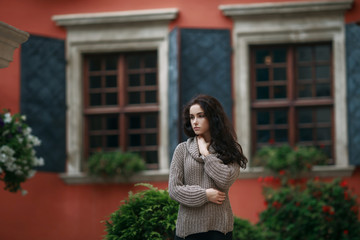 Beautiful young woman dressed in gray sweater staying in the city on a sunny day. Old town green garden and ancient windows background. Copy space