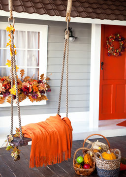 Veranda of countryside house in autumn season. swing is adorned with autumn leaves and orange knitted plaid. Basket with pumpkins and autumn vegetables.