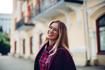 Cheerful pretty young lady holds his glasses her hands and smiles at his interlocutor.Girl wearing stylish burgundy coat and a shirt in a cage