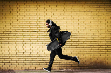 Fototapeta na wymiar Handsome young dreadlocks skater with headphones running in a suit near the yellow wall on the street.