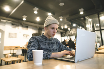 Fototapeta premium A hipster student sits in a cafe and works for a hand at a cup of coffee. Hipster sits in a cafe and uses a laptop.