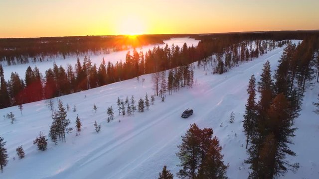 AERIAL: Flying above the car driving through snowy forest at golden winter sunrise. People on winter road trip traveling across snow covered Lapland wilderness at sunset. Car driving on empty icy road