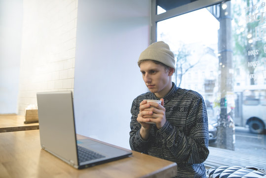 A young man is sitting in a cozy cafe with a cup of coffee in his hands and watching a video on a laptop. Stylish student at a computer in a cafe.