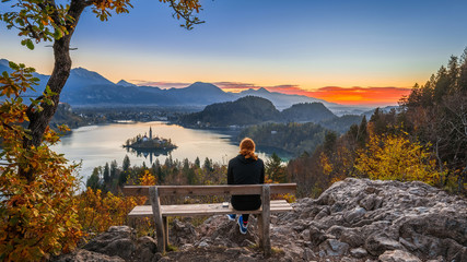Bled, Slovenia - Red hair runner woman enjoying the beautiful autumn view and the colorful sunrise of Lake Bled sitting on a hilltop bench wearing black pants and jumper with running shoes - Powered by Adobe