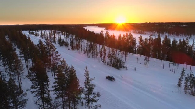 AERIAL: Flying above the car driving through snowy forest at golden winter sunrise. People on winter road trip traveling across snow covered Lapland wilderness at sunset. Car driving on empty icy road