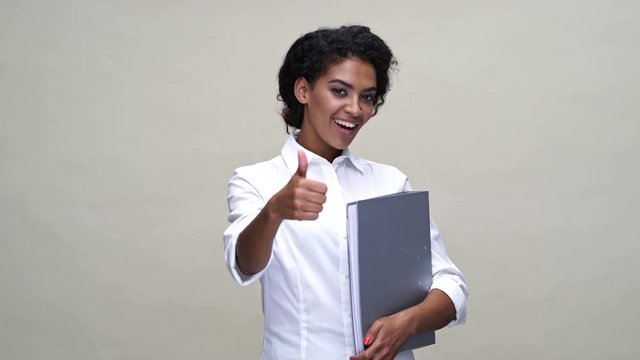 Happy african woman in shirt showing thumb up while holding folder with documents over gray background