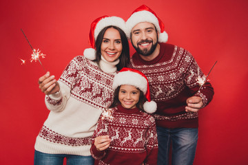 Fototapeta premium Cheerful relatives celebrate with sparkles, married couple, small cute adorable girl, in knitted traditional x mas costumes, head wear, beaming smiles, happiness