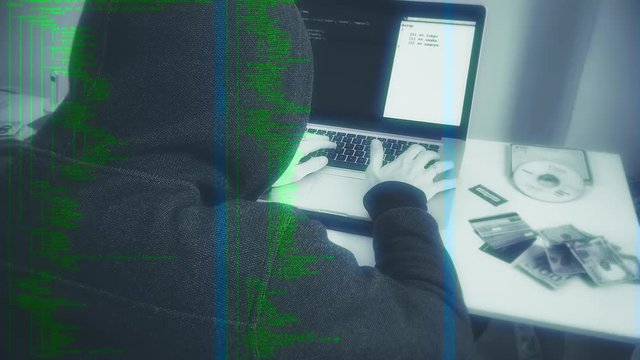 Hooded Hacker Coding On Computer, Slow Zoom. Hooded person typing in a laptop next to stolen itens, simulating a hacker crime scene