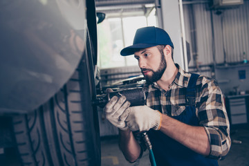 Serious brunet bearded brunet engineer handyman in checkered shirt, hat head wear is torquing lugs nuts of wheel at a garage station, ceiling background. Technology, safety concept