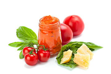 Transparent glass jar of red pesto with piece of cheese, cherry tomato and leaf of basil.