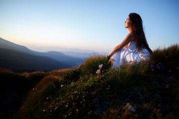 Beautiful woman in a long white dress in the mountains. Young woman sitting on a rock. Hair blowing in the wind. Girl relaxing outdoors. Beautiful woman posing in tall grass