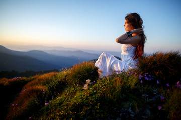 Beautiful woman in a long white dress in the mountains. Young woman sitting on a rock. Hair blowing in the wind