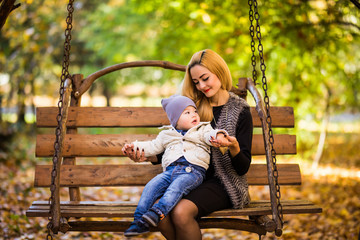 Young mother with her small son is resting on a wooden bench in the autumn golden park