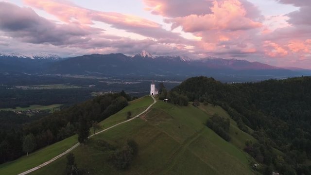 Colorful aerial view of the Jamnik Church on a hilltop at sunset while flying backwards and zooming into the landscape.