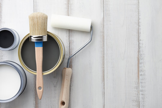 Paint brush, sponge roller, paints, waxes and other painting or decorating supplies on white wooden planks, top view