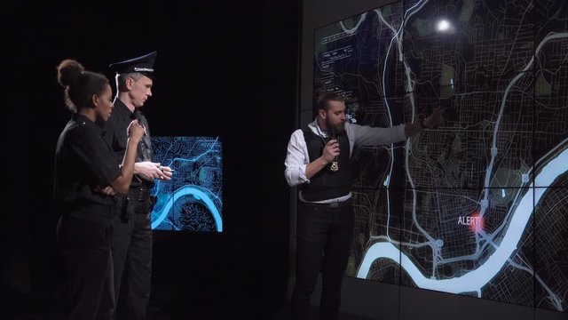 Coworking policemen watching GPS data on electronic map while spying bandit team and officers discuss response tactics in front of large live screens in a futuristic office.