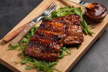 Closeup of pork ribs grilled with BBQ sauce and caramelized in honey on a bed of arugula. Tasty snack to beer on a wooden Board for filing on dark concrete background. Top view