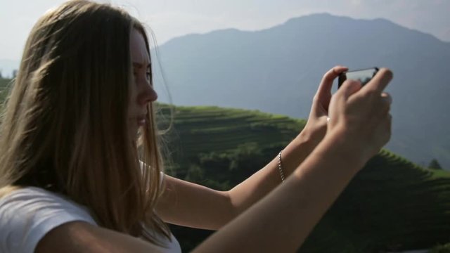 Young attractive woman watching sunrise over Longji Rice Terraces from top of the mountain. Blonde beautiful girl takes panorama photos on phone.