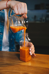Woman hand filling plastic bottle with fresh carrot juice on the wooden table in the kitchen. Close up