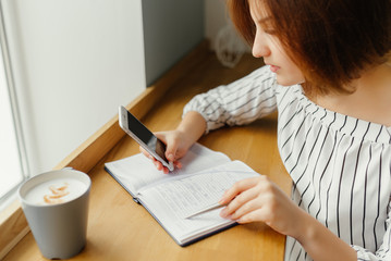 Naklejka premium Young business woman sitting in cafe at table and use smartphone and work with note book. Freelance work place.