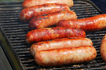 Bratwursts on a grill. Close up composition of grilled outdoor brat sausages during good weather 4th of July picnic. Lifestyle background, unhealthy eating concept.