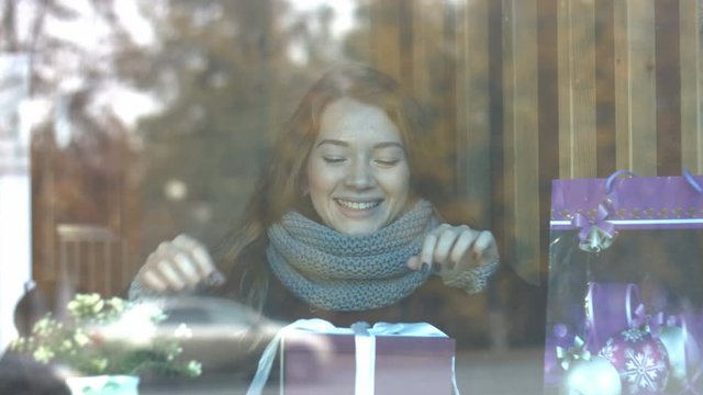 A girl with a gift in a cafe.
Slow motion. Young beautiful girl is sitting in a cafe and opens a box with a present gift.
She smiles. The glass reflects the traffic of urban transport.
