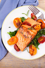 Salmon fillet with spinach and tomatoes on white plate on rustic wooden background. Overhead, top...