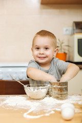 Little kid boy helps mother to cook Christmas ginger biscuit in light kitchen. Happy fair-haired child in gray T-shirt 2-3 years at the table with iron sieve and flour in weekend morning at home.