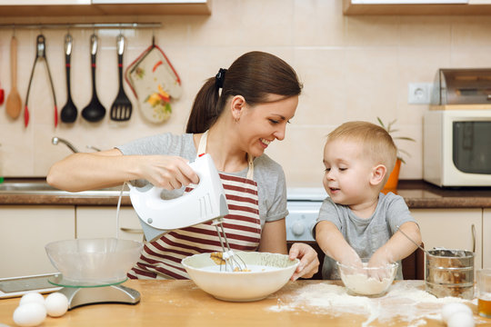 Little Kid Boy Helps Mother To Cook Christmas Ginger Biscuit In Light Kitchen With Tablet On The Table. Happy Family Mom 30-35 Years And Child 2-3 In Weekend Morning At Home. Relationship Concept