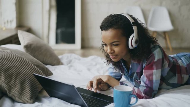 Young curly african american woman enjoying listen to music on laptop while lying on bed