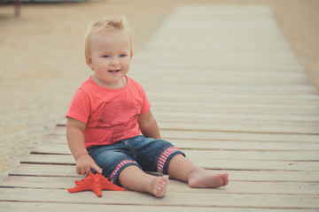 Naklejka premium Happy cute baby girl with blond hairs and blue eyes wearing stylish clothes posing smiling on sea side beach on wooden way with safety round and red sea star
