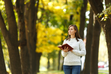 Naklejka premium A beautiful happy smiling brown-haired woman in white sweater standing with a red book in fall city park on a warm day. Autumn golden leaves. Reading concept.
