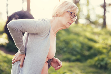 Problems with health. Unhappy pleasant aged woman holding her back and leaning forward while suffering from acute pain