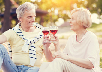 Day out. Positive nice elderly couple sitting together and drinking wine while having a picnic