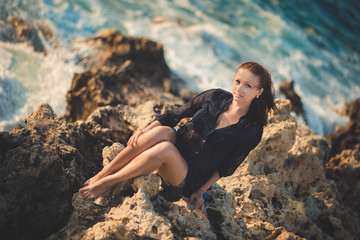 Hot sexy stylish dressed young lady brunette hairs and pinky cheeks with open legs shoulders and arms posing sensuality on sea side rocky beach. seductive woman awaiting her future seaman husband