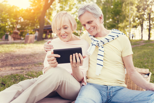 Modern Technology. Delighted Positive Elderly Couple Looking At The Tablet Screen And Smiling While Having A Great Time Together On A Picnic